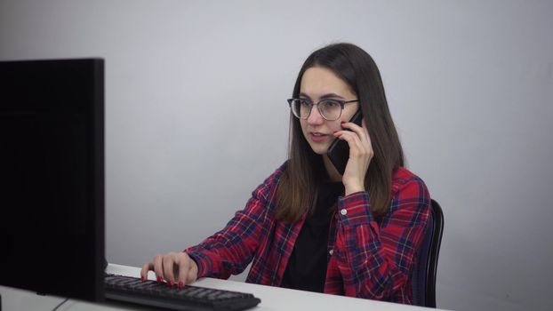 IT specialist sits at the workplace in the office and speaks on the phone. A young woman in glasses and a red shirt is working. 4k