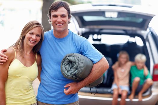 Couple ready to camp. Portrait of couple with man holding camping mattress and kids sitting in car