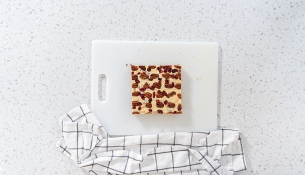 Flat lay. Cutting white chocolate cranberry pecan fudge into small pieces on a white cutting board.