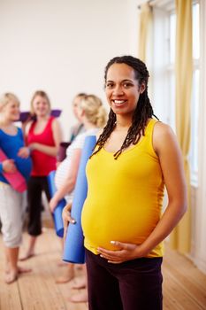Enjoying a healthy pregnancy lifestyle. A happy young pregnant African-American woman holding her exercise mat while standing in a gym with her friends in the background