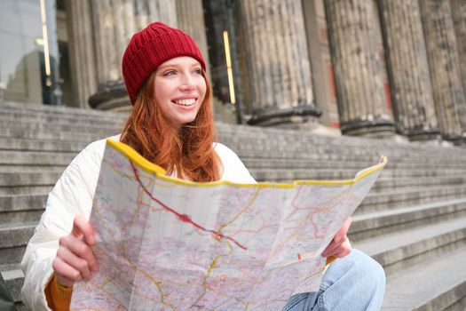 Portrait of young redhead woman, tourist sits with paper map and looks for a route to tourism attraction, rests on stairs outdoors.