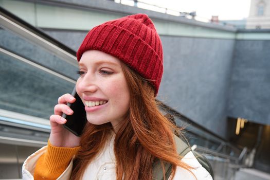 Portrait of happy redhead woman walking around town with smartphone, calling someone, talking on mobile phone outdoors.