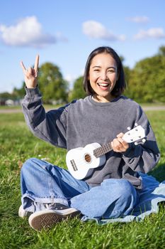 Happy asian girl playing ukulele in park, showing rock n roll, heavy metal horns sign and smiling, having fun outdoors.