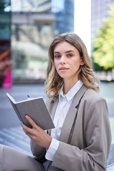 A young woman in corporate suit, sits with notebook and pen, takes notes, works and writes down her ideas.