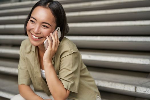 Close up portrait of smiling asian girl talks on mobile phone, sits outside on street stairs. Young woman calling friend on smartphone.