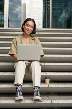 Young asian woman using laptop outdoors. Cute girl student with computer, does her homework, types, sits on stairs outside campus.