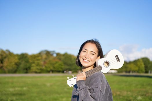 Young hipster girl, traveler holding her ukulele, playing outdoors in park and smiling. People concept