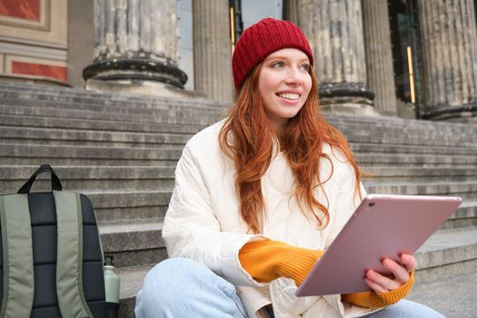 Beautiful young modern girl with red hair, holds digital tablet, sits on stairs near museum and connects public internet, sends message on gadget app.