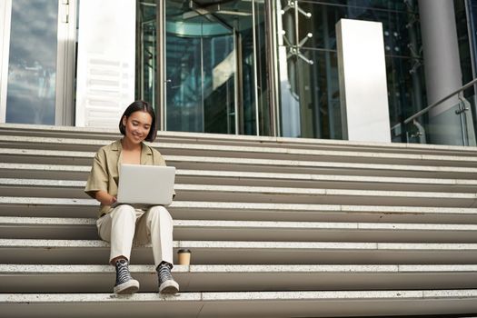 Asian girl student sits on stairs near campus, types on laptop, does her homework outdoors.