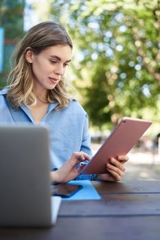 Vetical shot of young female student, reading on digital tablet, studying outdoors. Businesswoman sitting outside with laptop and gadgets, working.