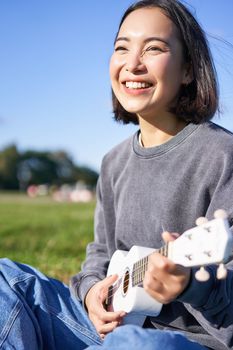 Vertical shot of happy korean girl sitting in park, learning how to play ukulele, singing and relaxing.