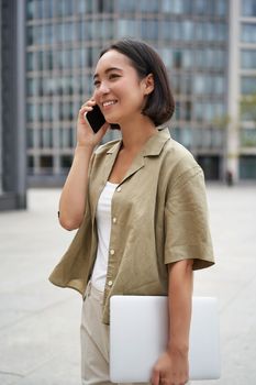 Vertical shot of young asian digital nomad, girl talks on mobile phone and walks on street with laptop. Young woman remote worker going to coworking space.