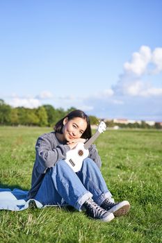 Romantic asian girl sitting with ukulele guitar in park and smiling, relaxing after university, enjoying day off on fresh air.