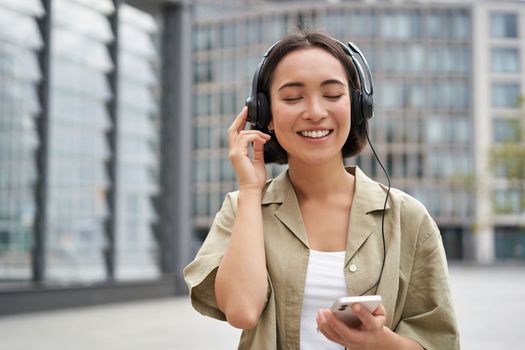 Streaming services concept. Happy asian girl listens music in headphones, holds mobile phone, choosing track or podcast, walking on street.