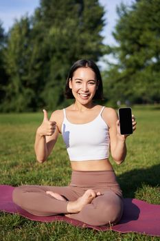 Vertical shot of asian woman shows thumbs up, recommending yoga training online, meditation app, doing exercises on fresh air in park, showing smartphone screen.