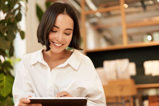 Portrait of asian girl drawing on digital tablet, scatching and doodling with graphic pen, sitting in cafe and smiling.