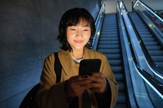 Happy smiling young woman, standing on escalator, going down, holding smartphone in both hands, chatting on mobile phone app.