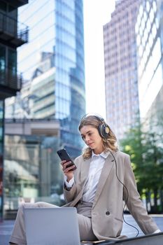 Corporate woman in suit, sitting in city, listening to music with laptop, scrolling news on her mobile phone, relaxing on lunch break.