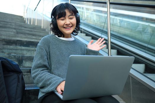 Smiling young student, wears headphones, sits on street stairs and waves hand at her laptop, connects to video chat, conference or training session on remote.