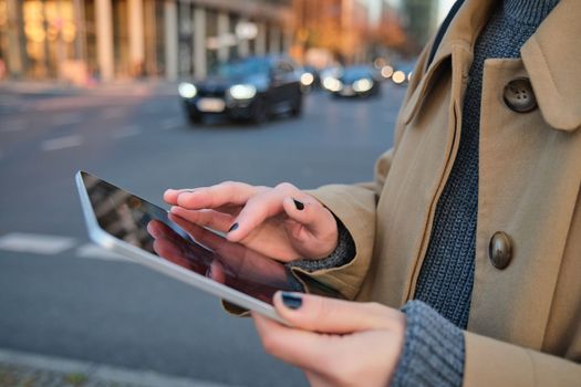 Close up photo of female hands holding digital tablet, girl looks at screen, checks application, stands on busy street filled with cars in afternoon.