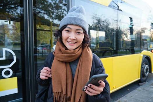 Image of girl student waiting for public transport, checks schedule on smartphone app, stands near city bus. Urban lifestyle concept