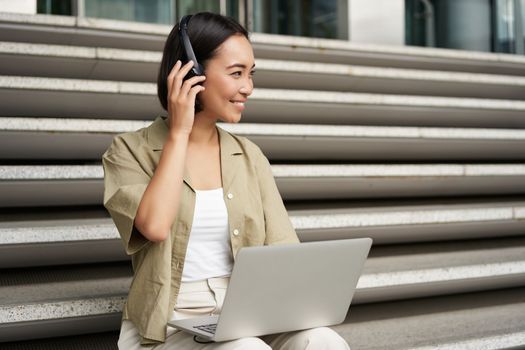 Portrait of stylish young woman smiles, sits on stairs with headphones and laptop, works on homework for university.