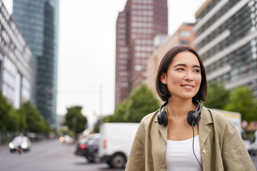 Portrait of young asian woman in headphones, posing in city, smiling and looking away, standing on street of city centre.