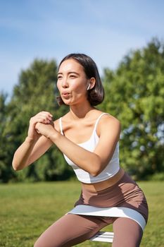Asian fitness girl doing squats in park, using resistance band, stretching yoga rope for workout training on fresh air.