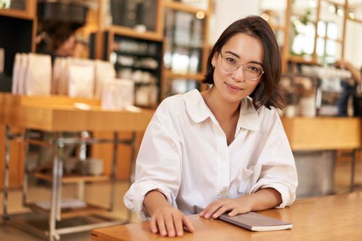 Portrait of brunette woman in cafe, sitting with notebook, working documents, looking at camera and smiling.