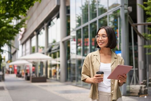 Smiling korean girl walking in city street with tablet, drinking takeaway coffee and looking happy.