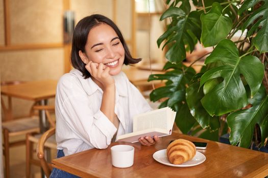 Romantic asian woman sitting with book in cafe, eating croissant and drinking coffee, reading and smiling, enjoying alone time.