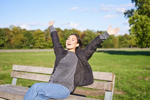 Happy asian woman stretching her hands, sitting on bench with excited face, smiling pleased, feeling freedom, enjoying day in park.