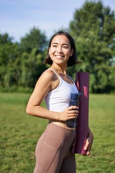 Vertical shot of fit and healthy asian woman posing in park, holding water bottle and yoga rubber mat for workout outdoors.