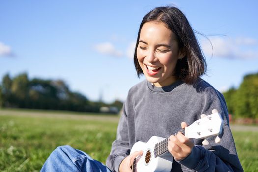 Happy people and hobbies. Smiling asian girl playing ukulele guitar and singing, sitting in park outdoors on blanket.