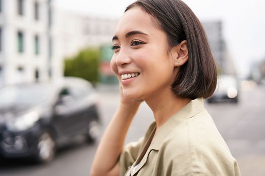 Close up portrait of beautiful young woman, asian girl stands outdoors on street, smiling happy.
