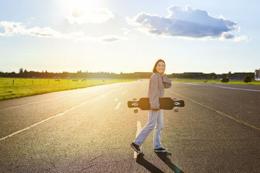 Young skater girl, teenager skating on cruiser, holding longboard and walking on concrete empty road.