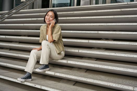 Portrait of asian student girl on stairs talks on mobile phone, smiles at camera, sits outside building.