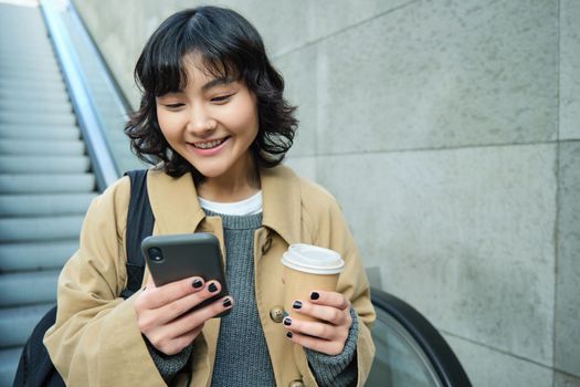 Portrait of smiling korean girl commutes, goes somewhere in city, drinks coffee to go and uses smartphone, stands on escalator.