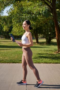 Active asian girl, in fitness clothing, workout in park, walking in sportswear with smartphone and water bottle.