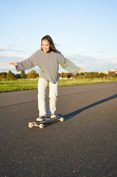 Cute asian girl riding skateboard, skating on road and smiling. Skater on cruiser longboard enjoying outdoors on sunny day.