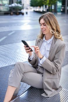 Vertical portrait of businesswoman in suit using her phone, relaxing outdoors, sitting on bench.