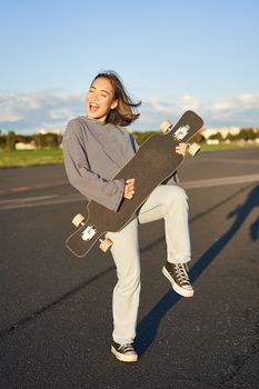 Funny asian girl enjoying skating, holding skateboard like guitar and shadow playing, having fun outdoors.