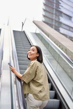 Young happy woman standing on escalator with smartphone, going up, walking in city.