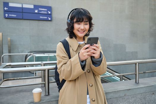 Portrait of stylish asian hipster girl, listens music in headphones, looks at her phone, tourist looking at map for a way to sightseeing, drinking coffee to go.