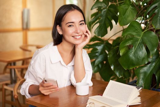 Beautiful young woman with smartphone, drinking coffee in cafe and smiling, relaxing with favorite book.