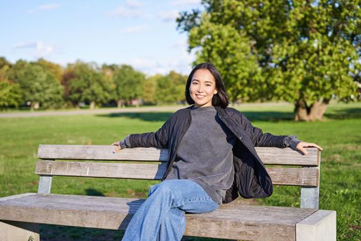 Smiling young asian woman in outdoor jacket, sitting on bench in green sunny park, resting alone, relaxing on fresh air.