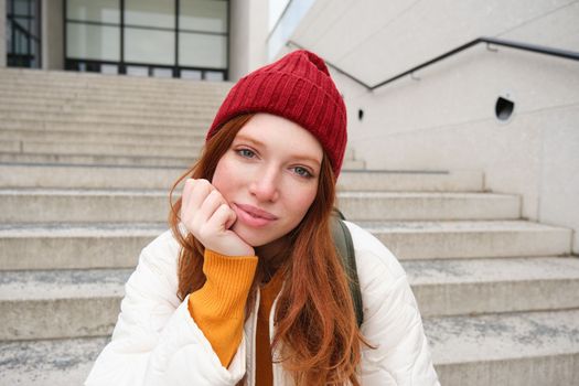 Close up portrait of beautiful redhead girl in red hat, urban woman with freckles and ginger hair, sits on stairs on street, smiles and looks gorgeous.