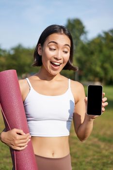 Excited fitness girl recommends application for sport and workout, shows phone screen, standing with rubber yoga mat in park after training session.
