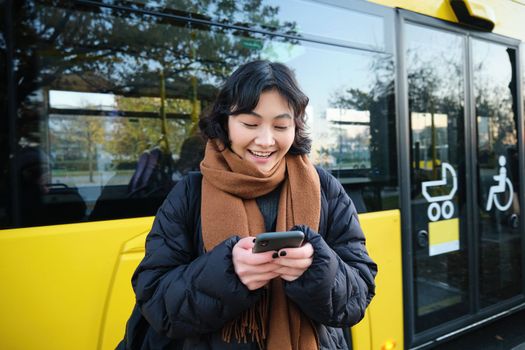 Beautiful korean girl, student on bus stop, looking at her smartphone, checking timetable, reading text message, wearing winter clothes.