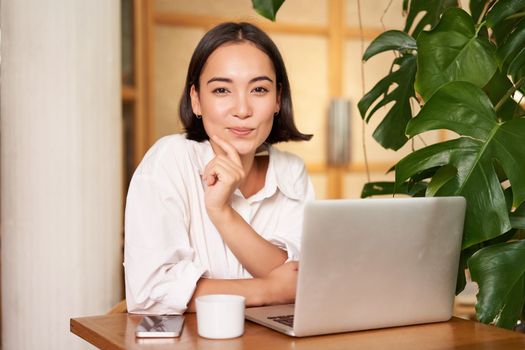 Confident young stylish woman with laptop, sitting in cafe and working, freelancer in co-working space with cup of coffee.